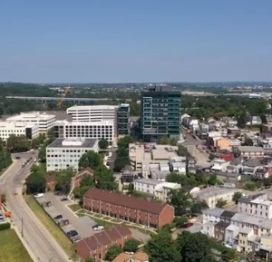 Aerial urban landscape with modern buildings, tree-lined roads, and green hills.