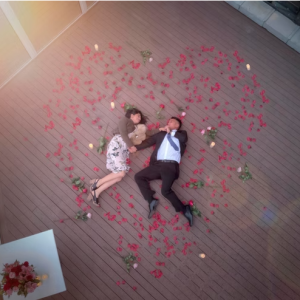 Romantic couple forming a heart amid red rose petals on a wooden deck.