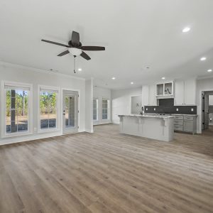Modern open-concept living room with kitchen island, wood floors, large windows, and ceiling fan.